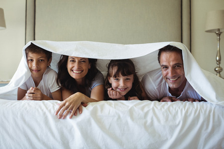 Portrait of happy family lying under blanket on bed in bedroom at homeの写真素材