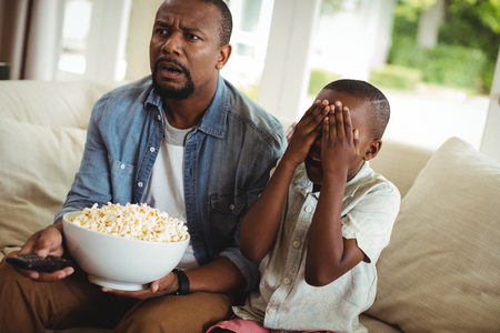 Son covering his eyes while watching television with father at homeの写真素材