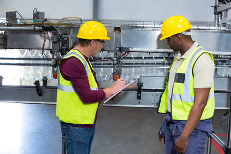 Two factory workers with clipboard working next to production line in drinks production plant