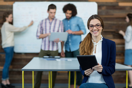 Portrait of smiling female business executive with digital tablet sitting in office and colleague...