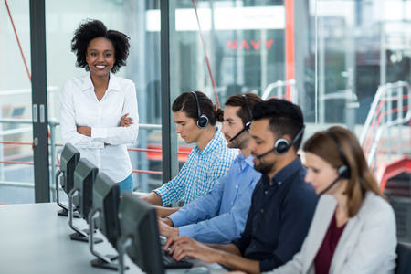 Smiling Businesswoman standing with arms crossed while colleagues talking on headset in office