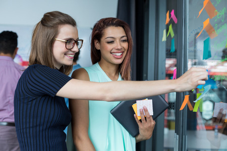 Smiling business executive sticking sticky note on glass in officeの写真素材