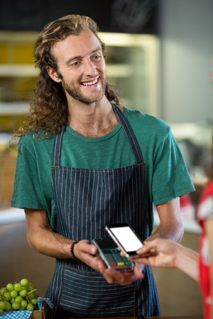 Smiling male staff receiving a payment through nfc technology at counter in shopの写真素材