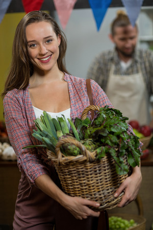 Portrait of smiling woman holding basket of green leafy vegetables in the grocery storeの写真素材