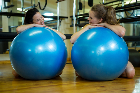 Portrait of happy women leaning on fitness ball in gymの写真素材