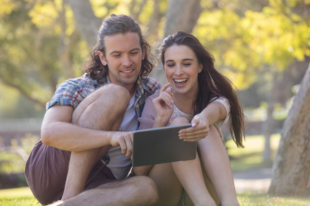 Couple sitting on grass and using digital tablet in park on a sunny dayの写真素材
