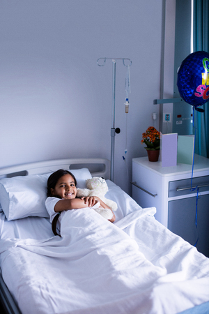 Portrait of smiling patient relaxing on bed with teddy bear in hospitalの写真素材