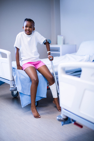 Girl with crutches sitting on hospital bed in ward of hospitalの写真素材