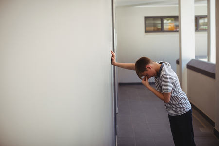 Sad schoolboy leaning against wall in corridor of schoolの写真素材