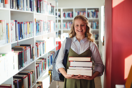 Portrait of schoolgirl standing with stack of books in library at school