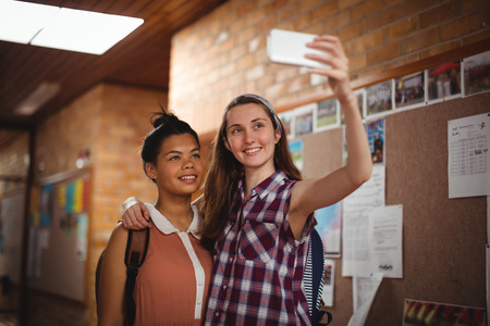 Happy schoolgirls taking selfie on mobile phone in corridor at school