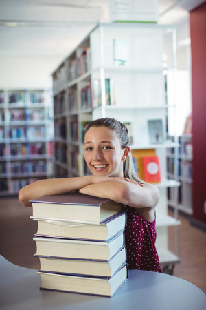 Portrait of happy schoolgirl leaning of stacked books in library at schoolの写真素材