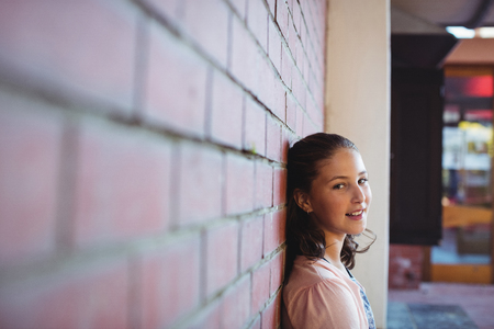 Portrait of happy schoolgirl sitting against brick wall in school campusの写真素材