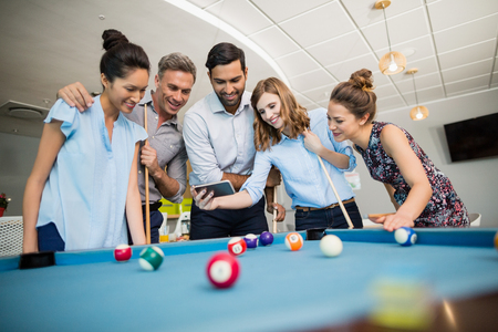 Smiling business colleagues using mobile phone while playing pool in office spaceの写真素材