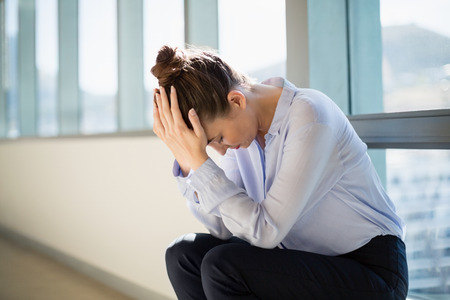 Tense female business executive sitting with hand on forehead in office corridor