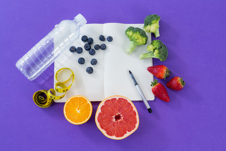Water bottle, measuring tape, various fruits, vegetable, opened book and pen on purple backgroundの写真素材