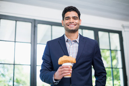 Portrait of businessman holding disposable coffee cup and croissant in a restaurantの写真素材