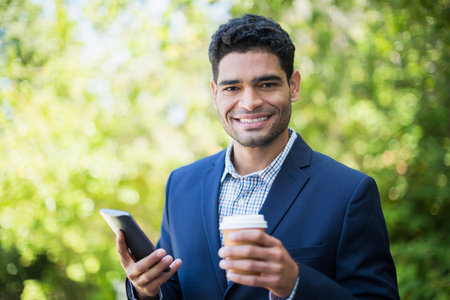 Portrait of businessman holding disposable coffee cup and mobile phone in a parkの写真素材