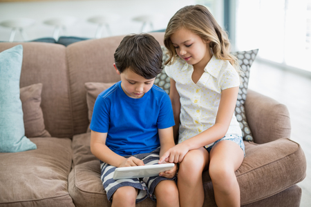 Siblings using digital tablet on sofa in living room at homeの写真素材