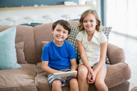 Portrait of smiling siblings using digital tablet on sofa in living room at homeの写真素材