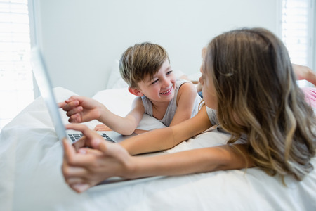 Smiling girl and boy using laptop on bed in bedroom at homeの写真素材