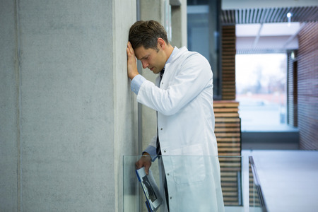Worried male doctor leaning on wall near corridor in hospitalの写真素材