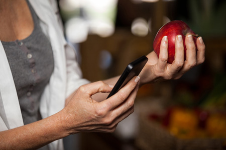 Mid section of female costumer holding apple while using mobile phone in supermarketの写真素材