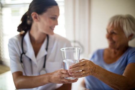 Female doctor serving a water glass to senior woman in the bedroom at homeの写真素材