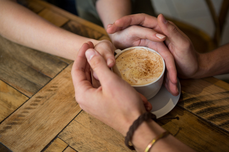 Close-up of couple holding hands and coffee cup in cafeteriaの写真素材