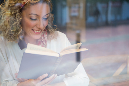 Smiling young woman reading book seen through cafe windowの写真素材
