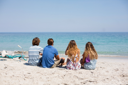 Rear view of friends sitting on shore at beach during sunny dayの写真素材