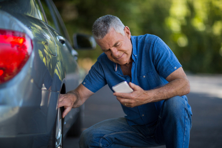 Senior man using phone while checking wheels of carの写真素材