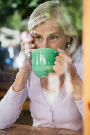 Senior woman drinking coffee while talking on mobile phone in cafe shopの写真素材