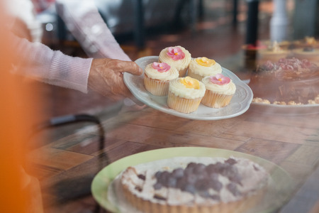 Close up of senior woman holding cupcakes in plate seen through glass windowの写真素材