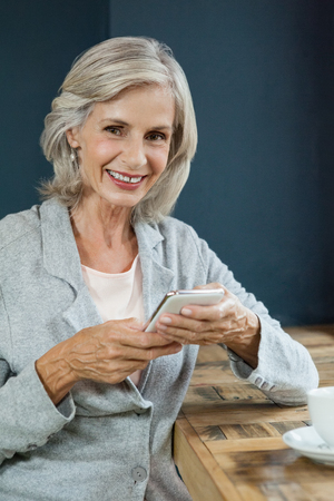 Portrait of smiling senior woman holding mobile phone while sitting at table in cafe shopの写真素材