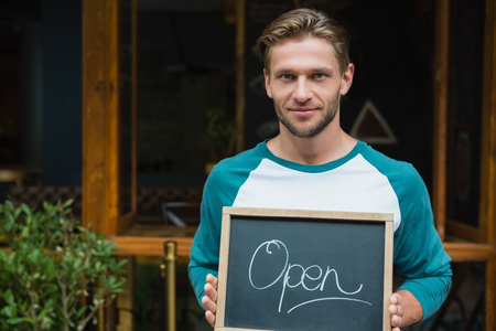 Portrait of smiling waiter holding chalkboard with open sign outside the cafeの写真素材