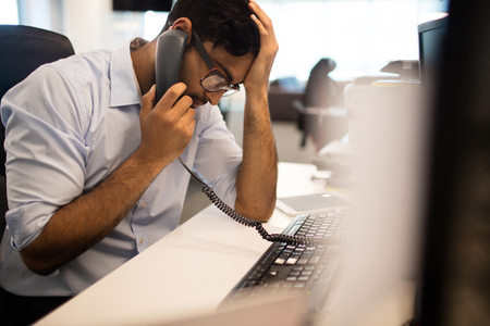 Tensed businessman with head in hand talking on land line in officeの写真素材
