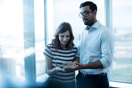 Business couple using mobile phone against glass window in office