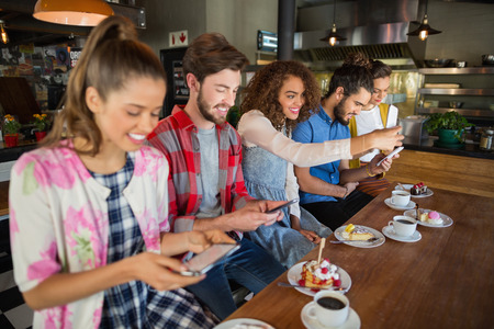 Smiling friends using their mobile phones while sitting by coffee cups and desserts on table in restaurantの写真素材