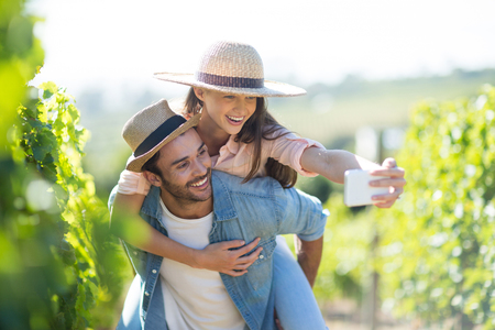 Happy couple taking selfie while piggybacking at vineyard during sunny dayの写真素材