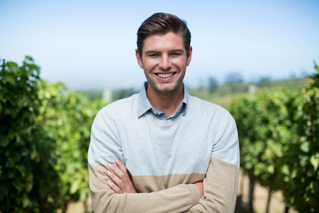 Portrait of smiling young man with arms crossed standing at vineyard against blue sky
