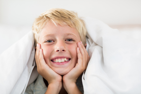 Portrait of smiling boy lying under bed sheet in bedroom at homeの写真素材
