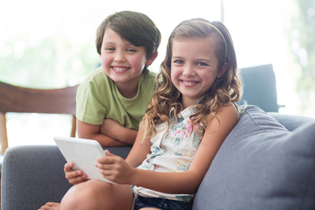 Portrait of siblings with digital tablet sitting on sofa in living room at homeの写真素材