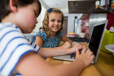 Smiling siblings using laptop in kitchen at home