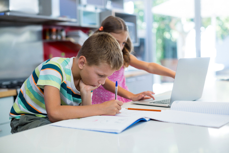 Boy doing his homework while girl using laptop in kitchen at homeの写真素材