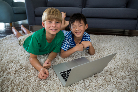 Portrait of smiling siblings with laptop lying on rug in living roomの写真素材