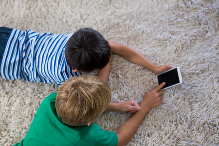 Siblings lying on rug and using mobile phone in living room at homeの写真素材