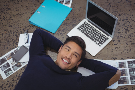 Smiling businessman lying by laptop and photographs on floor at officeの写真素材