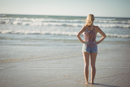 Rear view of woman standing with hands on hip at beach during sunny dayの写真素材