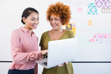Smiling businesswomen using laptop white standing against whiteboard in officeの写真素材